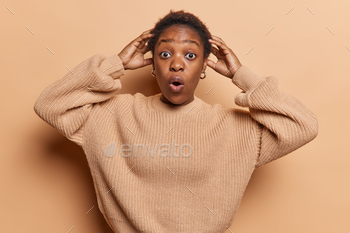 hair holds breath reacts to something amazing keeps hands on head has opened mouth dressed in knitted jumper isolated over brown background.