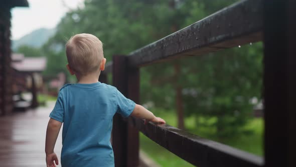Little Blond Child Walks Touching Wet Wooden Railing on Deck