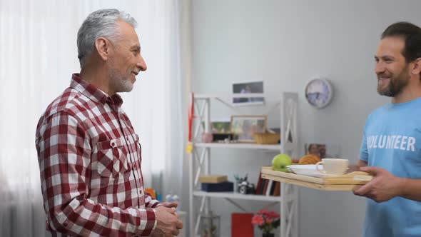 Middle Aged Volunteer Bringing Senior Man Tray With Breakfast, Looking to Camera