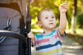 Pensive toddler boy stands near the stroller in the park with his hand raised.  - PhotoDune Item for Sale