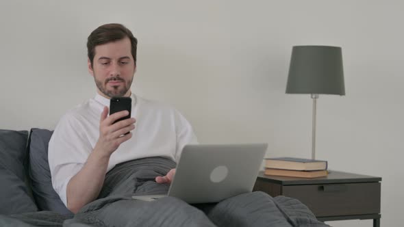 Young Man with Laptop Using Smartphone in Bed