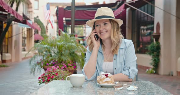 Positive Smiling Woman Is Talking with Friend By Phone Sitting at Street Cafe