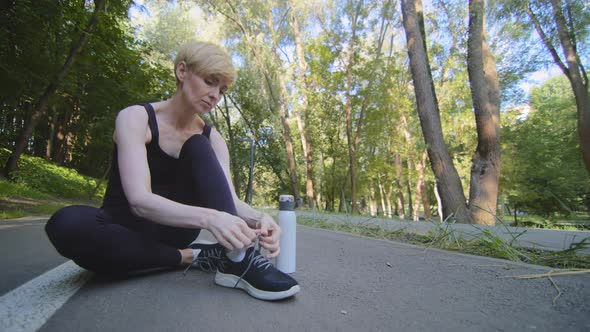 Middle Aged Caucasian Woman Athlete Runner Sitting on Track in Park Tying Laces on Sneakers Athletic