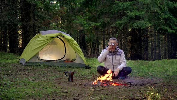 A Man Drinks Tea Near a Tent in the Forest