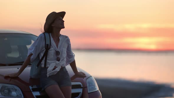 Pleasant Young Travel Woman Relaxing Waving Hair Sitting on Car Bonnet Enjoying Dusk Sunset Seascape