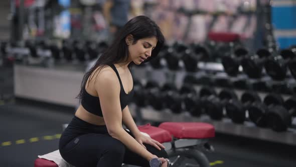 Confident Middle Eastern Woman Sitting on Exercise Bench in Gym Lifting Dumbbell