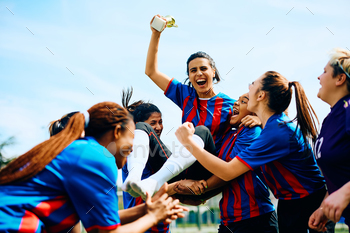 Cheerful soccer plyer holding the winning trophy while being carried by her teammates on stadium.