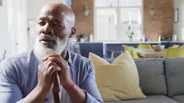 Thoughtful senior african american man in living room sitting on sofa, holding walking cane