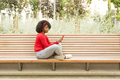 Smiling young afro woman on park bench using phone and headphones to listen to music. - PhotoDune Item for Sale