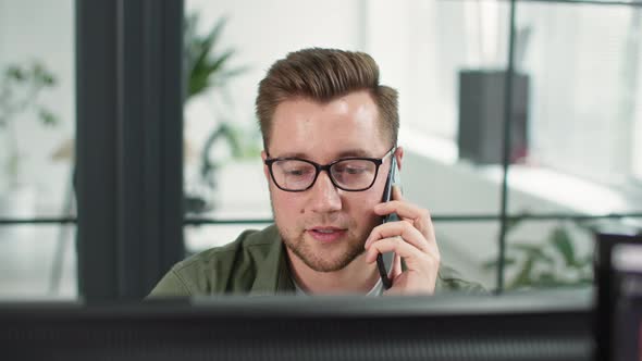 Smiling Male Employee Wearing Glasses Talks to Client on Phone While Working at Computer or