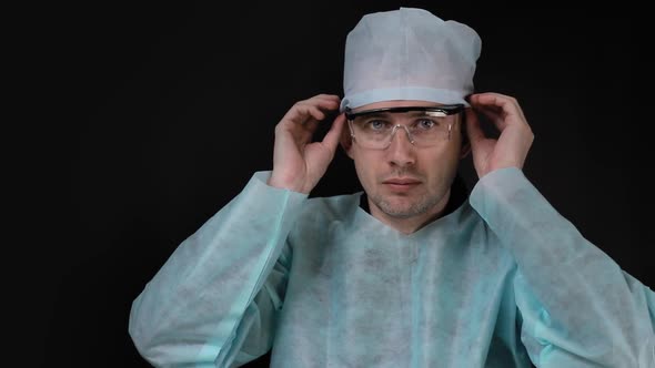 Portrait of a Doctor Surgeon Who Sits at a Table and Adjusts His Hat and Goggles on a Black