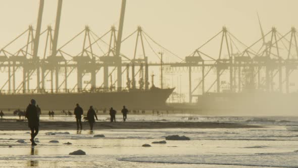 People silhouettes walk on beach in sunset with industrial port crane backdrop