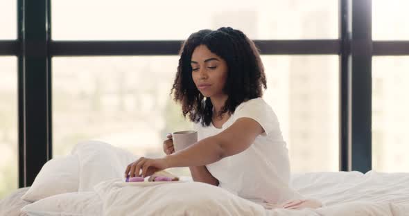 Young African American Lady Enjoying Breakfast in Bed, Eating Cookies with Coffee in Morning