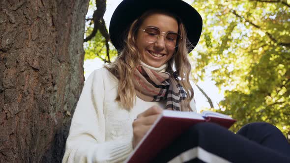 Cute Blonde Woman in the Hat Sitting Near Tree in the Park