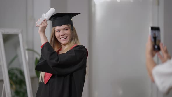 Excited Graduate Daughter Posing with Rolled Diploma As Mother Taking Photos on Smartphone Indoors