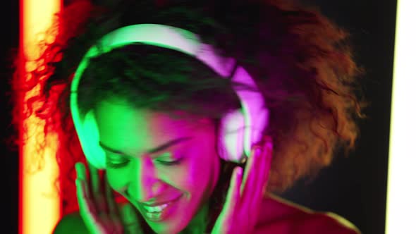 Mixed Race Woman with Afro Hairstyle Dancing in Wireless Headphones in Room with Colorful Neon Lamps
