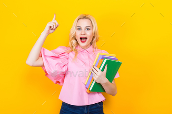 uth in pink shirt and jeans with pile of books isolated on orange background with copyspace and pointing with finger up, coming up with idea concept.