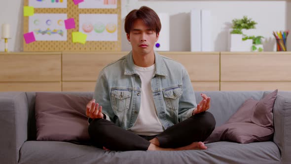 Clam of Asian young man doing yoga lotus pose to meditation and relax on couch during work online at