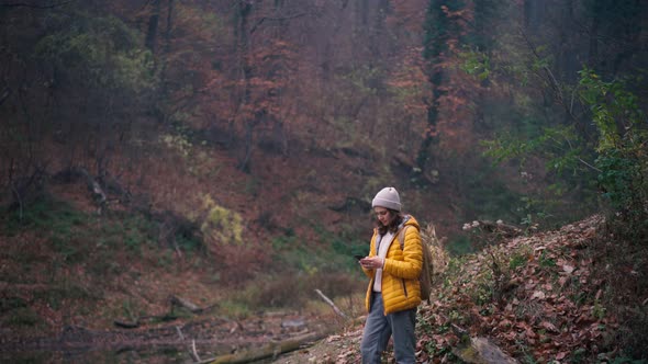 A Young Woman with a Backpack Using Her Phone While Standing at the Forest Lake