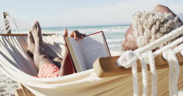 Senior african american man reading and lying in hammock on sunny beach