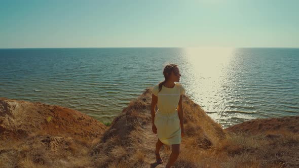 Happy Smiling Woman Traveler in Yellow Dress Walks on Edge of Clay Hill Over Sea with Morning