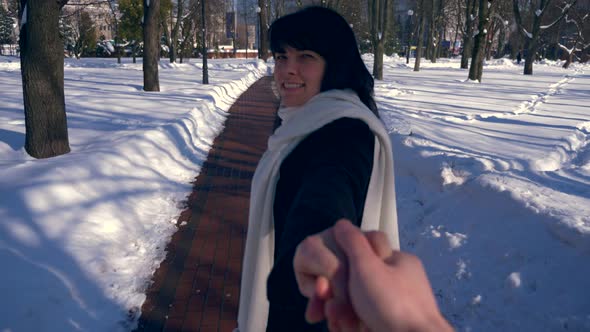 Pretty Smiling Girl Holds Man Hand. Follow Walking In Snow Covered Park. Winter City