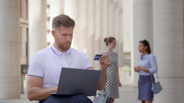 Medium Shot of Caucasian Businessman Using Laptop and Credit Card Outdoors Office Building