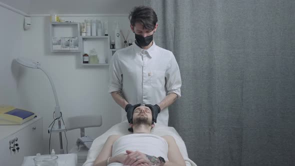 Portrait of Professional Confident Male Beautician in Coronavirus Face Mask Doing Facial Skin