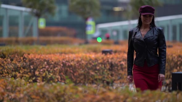 a Beautiful Business-like Confident Woman in a Purple Cap and Leather Jacket Strolls Along the City