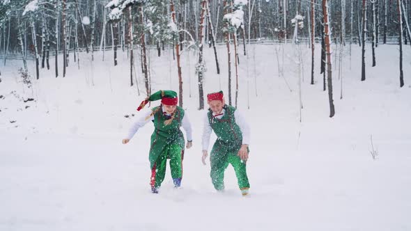 Two Elves in Green Suits with Hats are Playing and Throwing Up Snow with Their Hands 