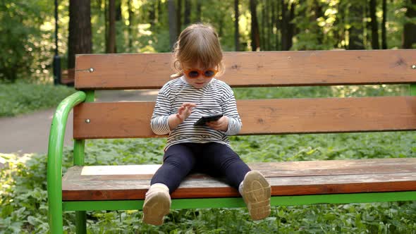 Beautiful Little Girl with Blond Hair and Ponytail in Orange Funny Glasses Dials Number on Phone and