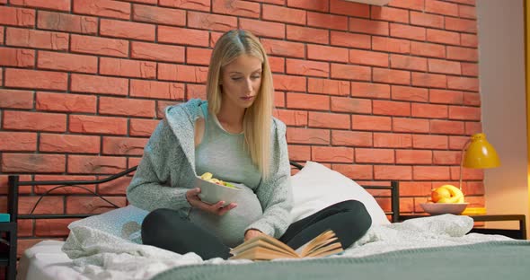 Beautiful Pregnant Woman is Reading Book Sitting in Bed at Home and Smiling Enjoying Literature