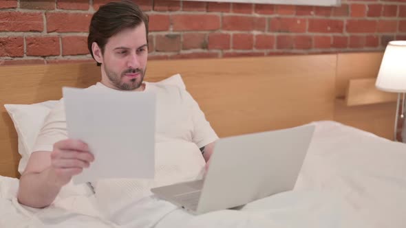 Casual Young Man with Laptop Having Loss on Documents in Bed