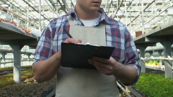 Professional Gardener in Apron Writing Notes on Clipboard Working in Greenhouse alt