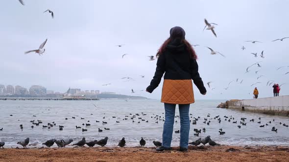 On the beach sand is a woman in a coat, and looks at the flying gulls in the sky. Rear view