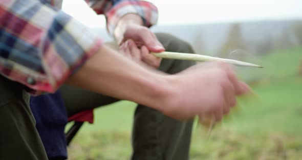 Close Up of Hands of Man Cutting with Knife a Sprig, Sitting on Camping Chair. Cloudy Sky in the