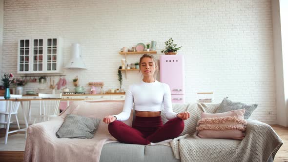 Beautiful Young Woman Meditating on Couch in Lotus Position with Bright Kitchen Background