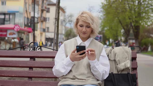 Young Blonde Business Woman in a Beige Vest and White Blouse Sits on a Bench in the City and Works