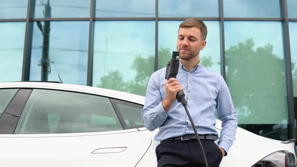 Hansome Guy Standing Near His New Modern Electric Car and Holding Plug of the Charger