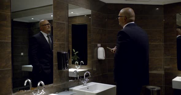 Serious Businessman Is Walking on Hall of Restroom in Hotel, Holding Smartphone