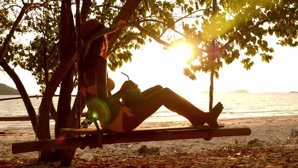 Beautiful woman in bikini sitting on wooden swing under tree on the beach