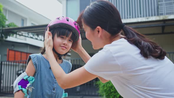 Asian loving mother put on sport helmet to little cute kid daughter before ride Surf Skate Board.