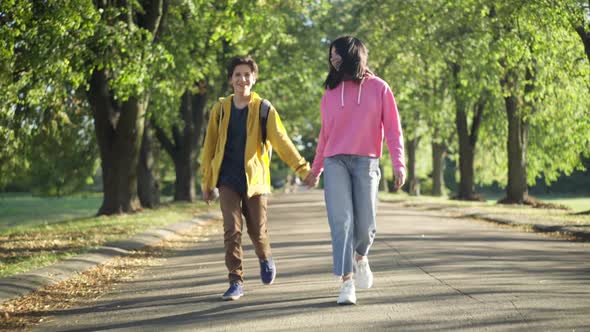 Dolly Shot of Happy Carefree Loving Teenage Couple Strolling in Sunshine on Park Alley Holding Hands