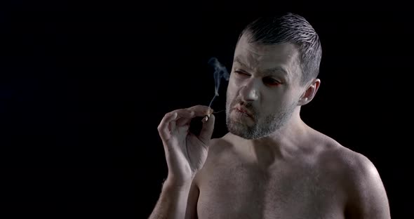 Aggressive Man with White Greasepaint on Face and Body Is Smoking Cigarette in Dark Room