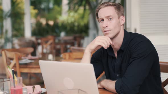 Businessman Sitting at Table with Laptop in Modern Cafe, Looks at Camera