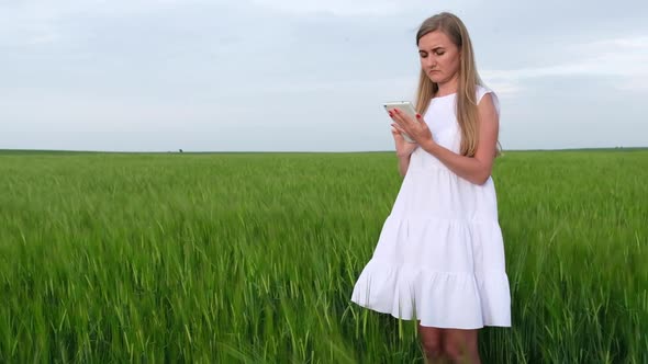 Young Girl in a White Dress with a Tablet in His Hands Stands in a Green Field of Wheat