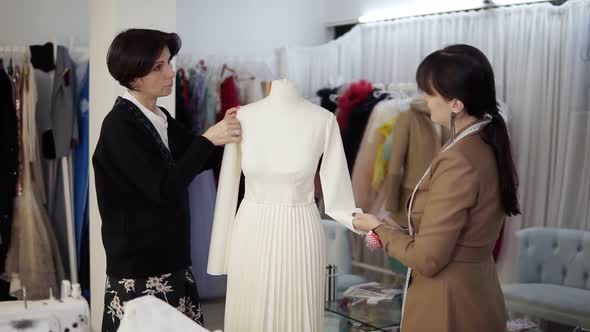 Two Caucasian Female Tailors Collaborating on White Wedding Dress and Pinning Unfinished Garment on