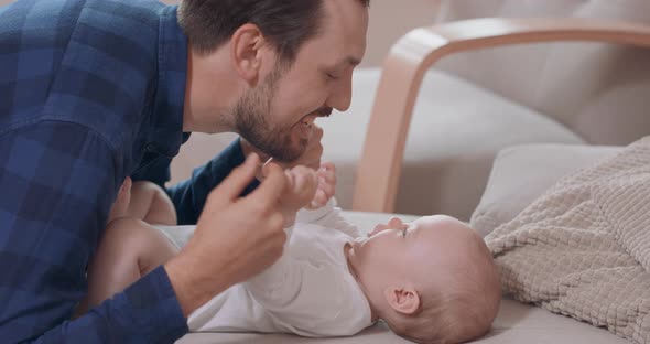 Close Up of Bearded and Mustashed Father is Kneeling By the Baby Lying on the Couch Plays with Its