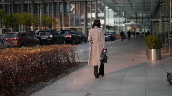 a Brunette in a Gray Coat and Highheeled Shoes Walks and Turns Around at a Modern Building at Dusk