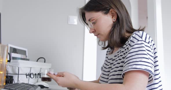 The young woman is putting on eyeglasses, holding smartphone looking at phone screen 